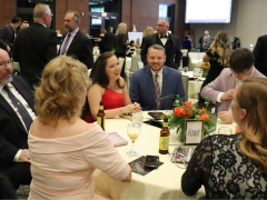 People in formal attire sit at a round table with drinks and programs at a banquet or gala event, with more guests and tables visible in the background.