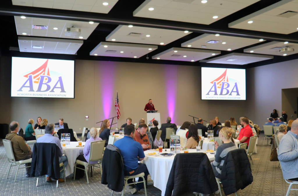 People seated at round tables in a conference room while a speaker presents at a podium. Two large screens display the AGrowth Business Association logo.