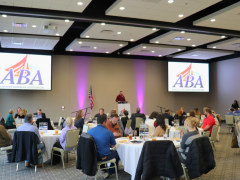 People seated at round tables in a conference room while a speaker presents at a podium. Two large screens display the AGrowth Business Association logo.