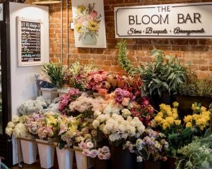 A flower shop interior with various fresh floral arrangements and bouquets displayed in vases below a sign reading “The Bloom Bar” on an exposed brick wall.