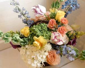 A mixed floral arrangement featuring white hydrangeas, yellow roses, pink carnations, purple-tipped tulips, blue delphiniums, and green foliage in a basket.