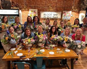 A group of women smile and hold floral arrangements while gathered around a wooden table at a flower workshop inside The Bloom Bar.