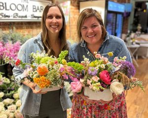 Two women standing indoors, each holding a large, colorful floral arrangement and smiling, with a flower shop setting in the background.