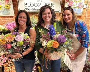 Three women stand indoors holding colorful flower arrangements, smiling at the camera. A sign behind them reads "The Bloom Bar." Shelves of flowers are visible in the background.
