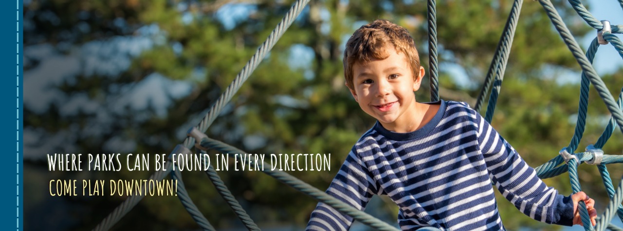 Young boy wearing a striped shirt climbs on rope playground equipment outdoors, with trees blurred in the background. Text on image promotes visiting downtown parks.