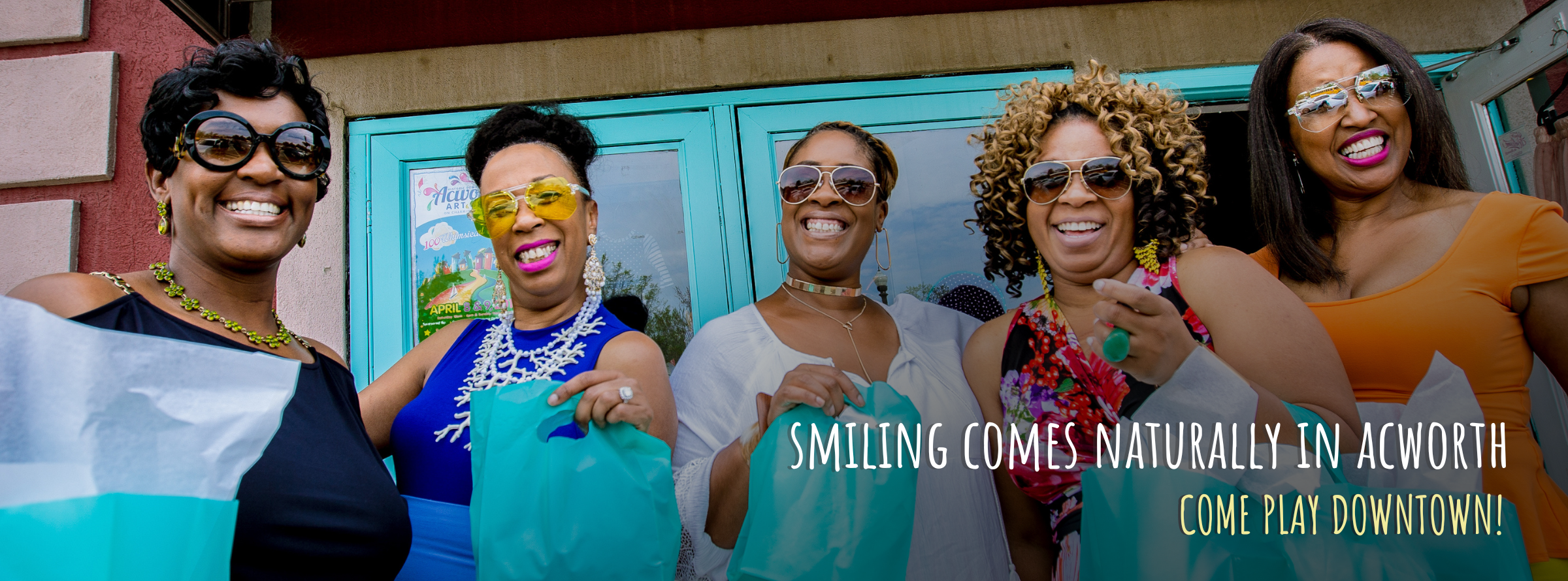 Five women stand outside a building, holding blue shopping bags and smiling at the camera. Text reads: "Smiling comes naturally in Acworth. Come play downtown!.