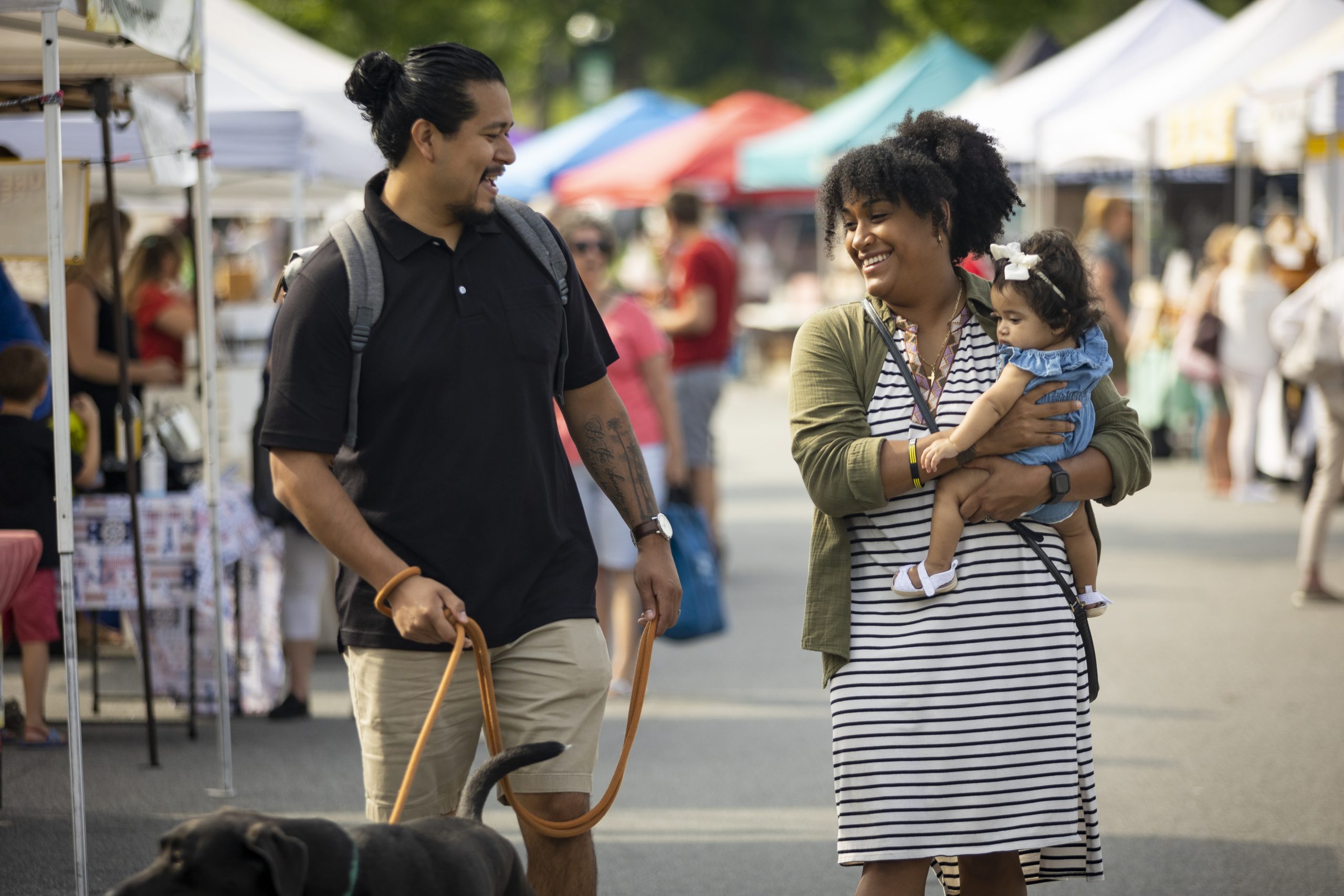 A man and woman walk outdoors at a market with a young child and a black dog, surrounded by vendor tents and other people.