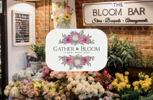 A flower shop interior with assorted flowers, a chalkboard price list, and a sign reading "Gather & Bloom Floral Boutique" in front of exposed brick walls.