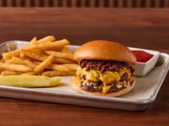 A cheeseburger with melted cheese and crispy onions on a bun, served with French fries, a pickle spear, and a side of ketchup on a metal tray.
