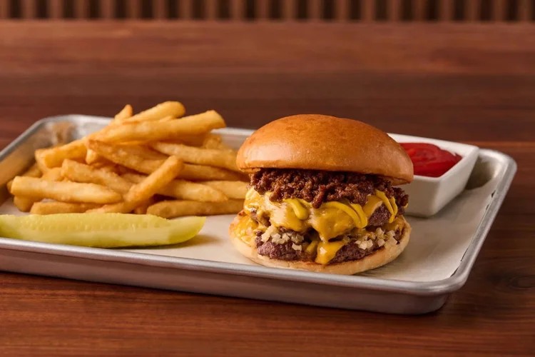 A cheeseburger with melted cheese and crispy onions on a bun, served with French fries, a pickle spear, and a side of ketchup on a metal tray.