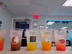 Five colorful drinks in clear plastic cups labeled "Dockside Nutrition," each with different stickers, are lined up on a counter in a brightly lit indoor setting.