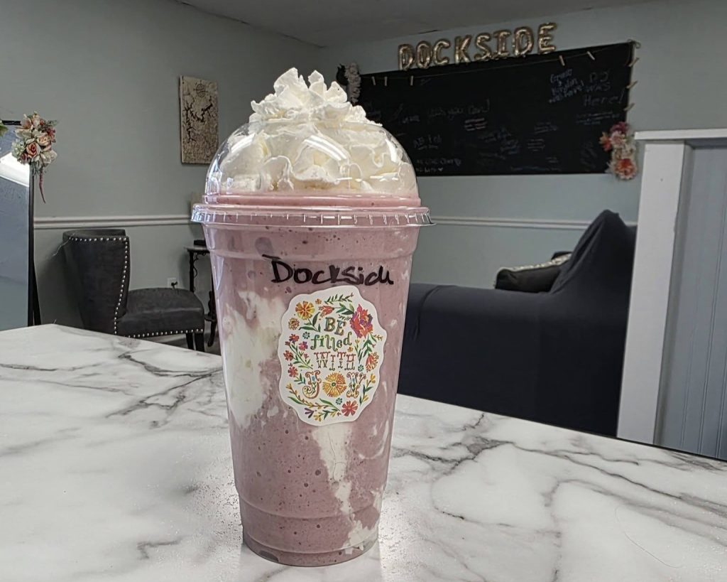 A plastic cup filled with a pink and white blended drink topped with whipped cream sits on a marble countertop in a cafe setting.