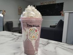 A plastic cup filled with a pink and white blended drink topped with whipped cream sits on a marble countertop in a cafe setting.