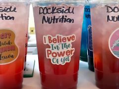A clear plastic cup labeled "Dockside Nutrition" filled with a red and pink drink; the cup features a sticker that reads "I Believe In The Power Of Yet." Other similar drinks are nearby.