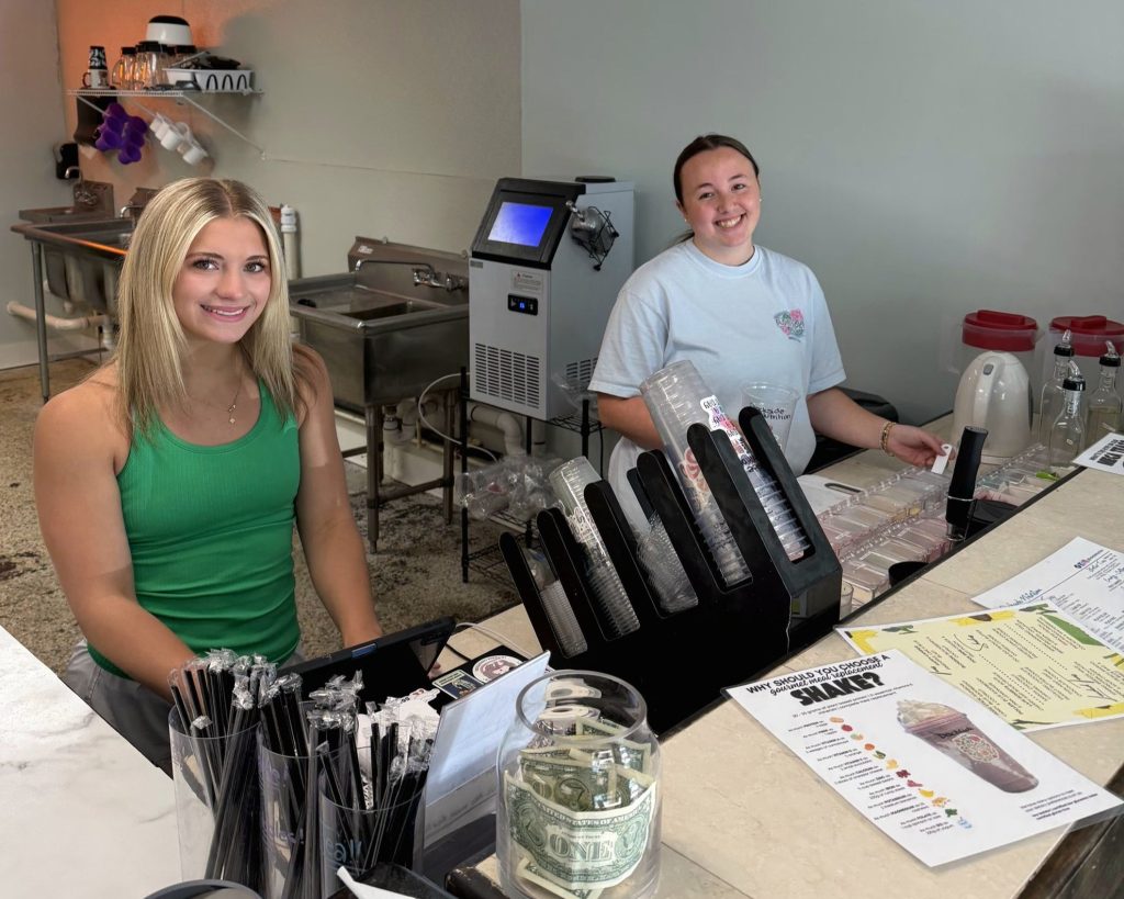 Two women stand behind a counter at a café, smiling. Menus, straws, a tip jar with cash, and drink machines are visible on the counter and in the background.