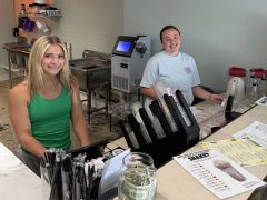 Two women stand behind a counter at a café, smiling. Menus, straws, a tip jar with cash, and drink machines are visible on the counter and in the background.