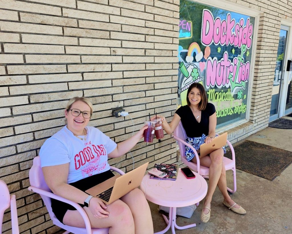 Two women sitting at a pink table outside a nutrition shop, working on laptops and clinking drinks, with a colorful shop sign in the background.