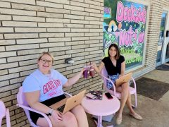Two women sitting at a pink table outside a nutrition shop, working on laptops and clinking drinks, with a colorful shop sign in the background.