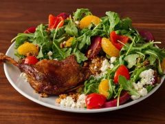 A plate with roasted duck leg, arugula salad, mandarin orange slices, cherry tomatoes, red peppers, beets, crumbled cheese, and granola on a wooden table.