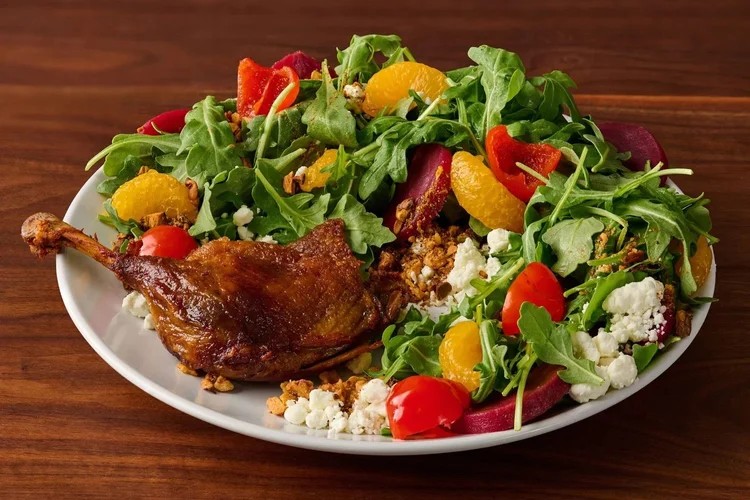 A plate with roasted duck leg, arugula salad, mandarin orange slices, cherry tomatoes, red peppers, beets, crumbled cheese, and granola on a wooden table.