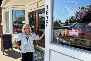 A woman stands smiling at the entrance of Pearl's Spa, next to a glass door and storefront window displaying the spa's name, street number 4827, and a flyer for Café de Coco.