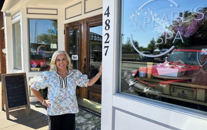A woman stands smiling at the entrance of Pearl's Spa, next to a glass door and storefront window displaying the spa's name, street number 4827, and a flyer for Café de Coco.