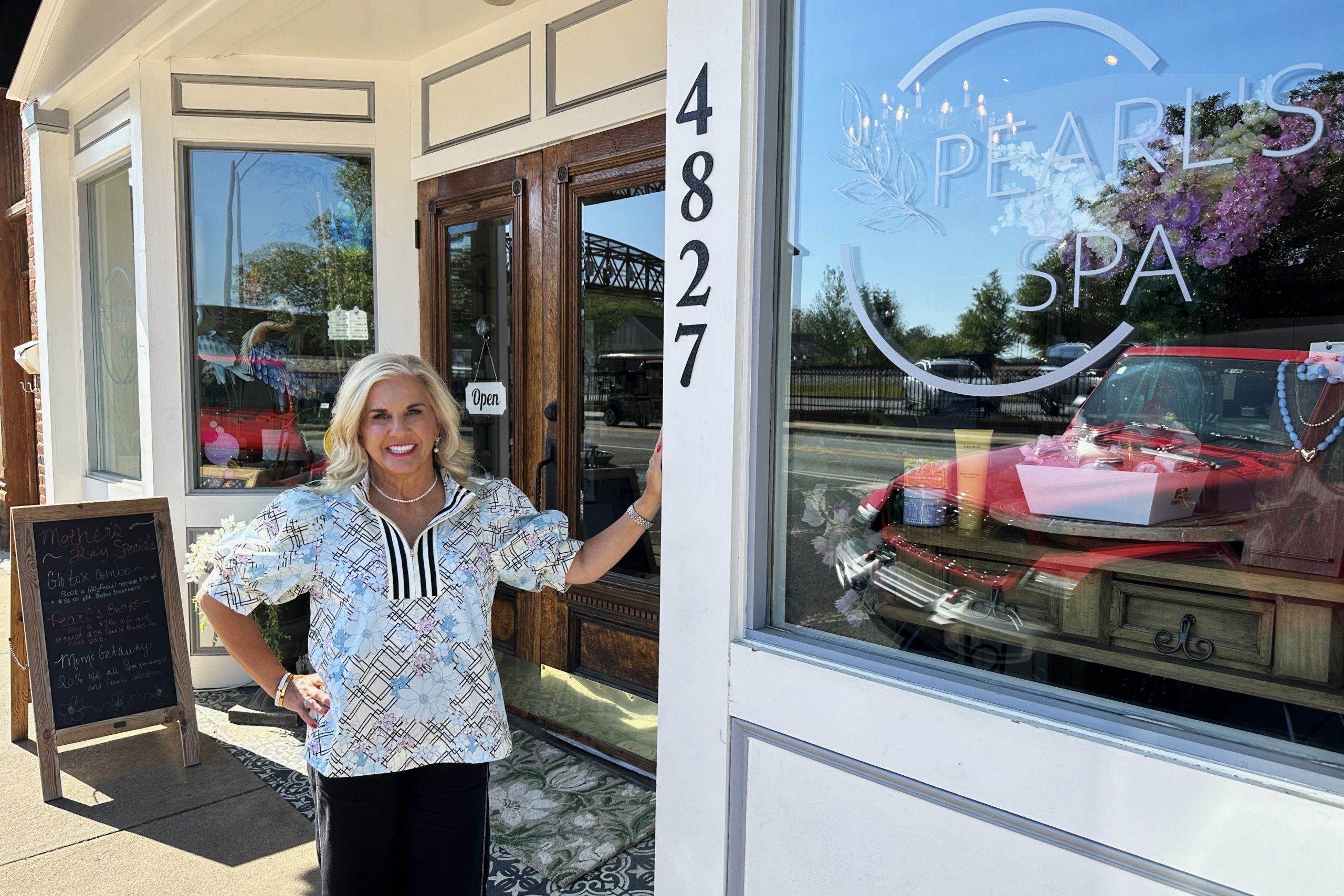 A woman stands smiling at the entrance of Pearl's Spa, next to a glass door and storefront window displaying the spa's name, street number 4827, and a flyer for Café de Coco.