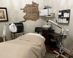 A treatment room at Pearl's Spa in Downtown Acworth features a beige-covered massage table, magnifying lamp, facial equipment, shelves with skincare products, and an exposed brick wall for a serene ambiance.