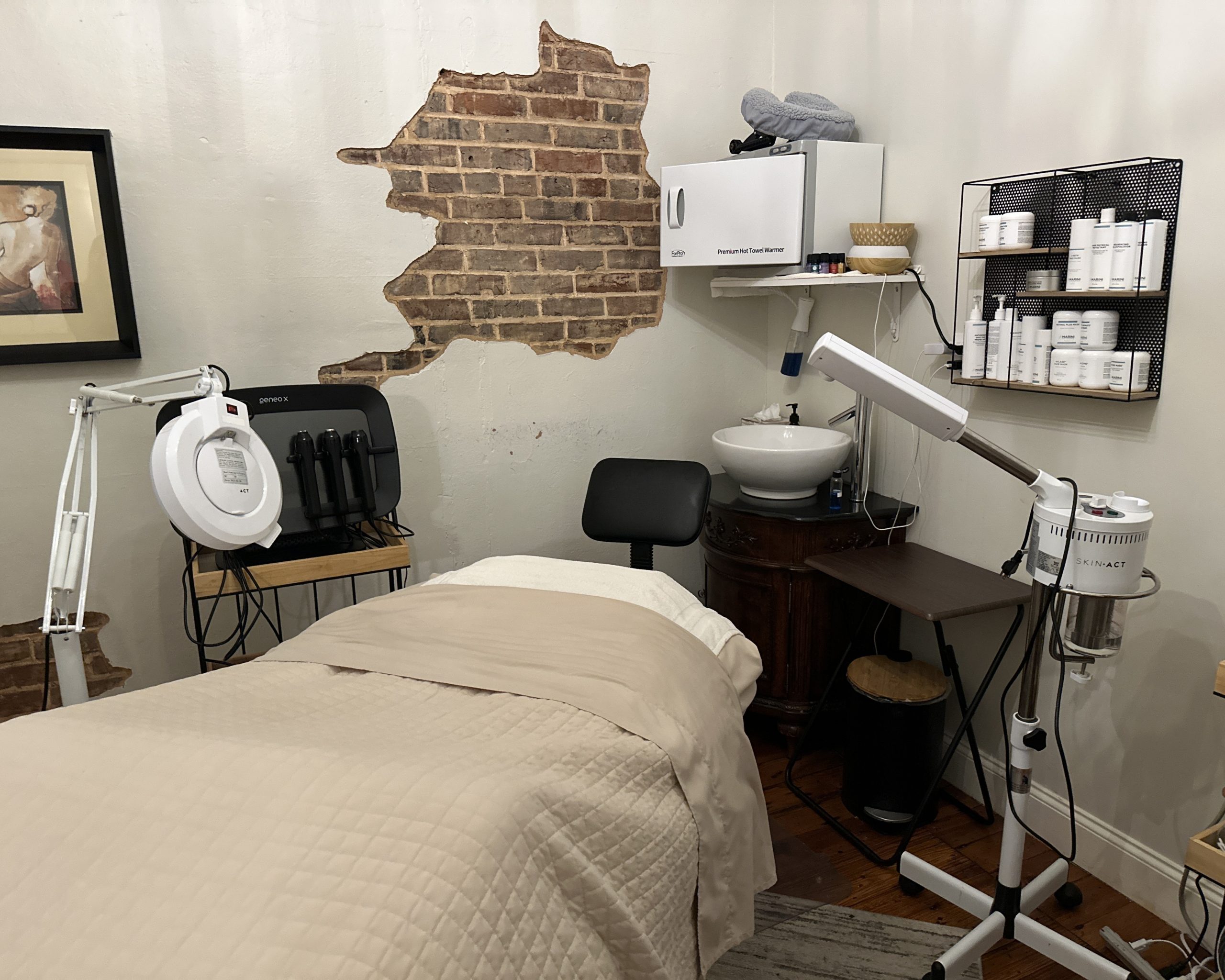 A treatment room at Pearl's Spa in Downtown Acworth features a beige-covered massage table, magnifying lamp, facial equipment, shelves with skincare products, and an exposed brick wall for a serene ambiance.