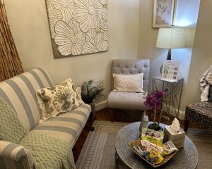 A cozy waiting room at Pearl's Spa in Downtown Acworth features a striped sofa, armchair, lamp, floral wall art, and a small table with magazines, snacks, tissues, and a plant in the corner.