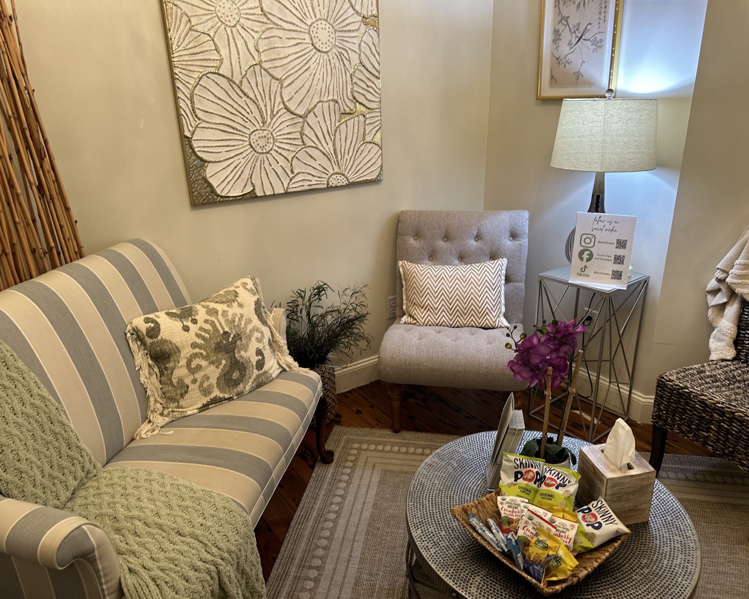 A cozy waiting room at Pearl's Spa in Downtown Acworth features a striped sofa, armchair, lamp, floral wall art, and a small table with magazines, snacks, tissues, and a plant in the corner.