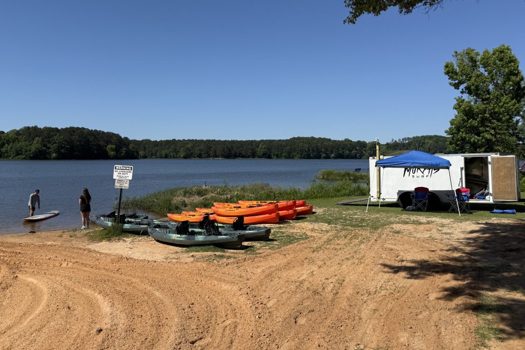 Kayaks and a paddleboard are lined up by a lake with a trailer and tent in the background.