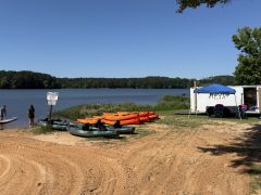 Kayaks and a paddleboard are lined up by a lake with a trailer and tent in the background.
