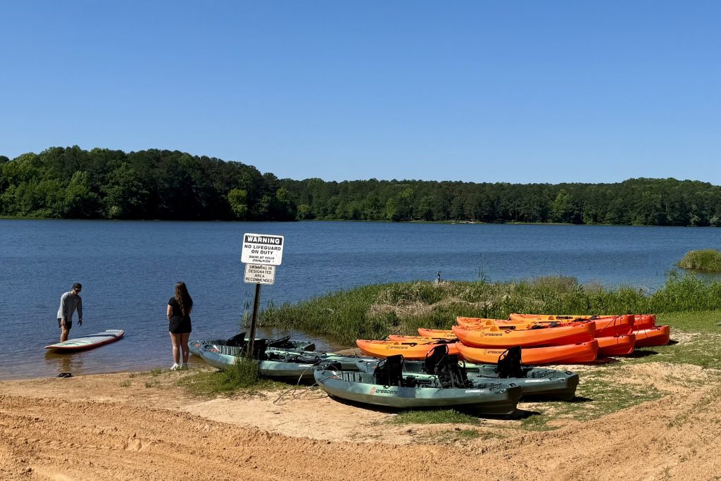Two people stand by the edge of a lake near kayaks and paddleboards on a sandy shore, with a warning sign posted nearby and trees in the background.