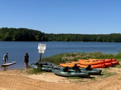 Two people stand by the edge of a lake near kayaks and paddleboards on a sandy shore, with a warning sign posted nearby and trees in the background.