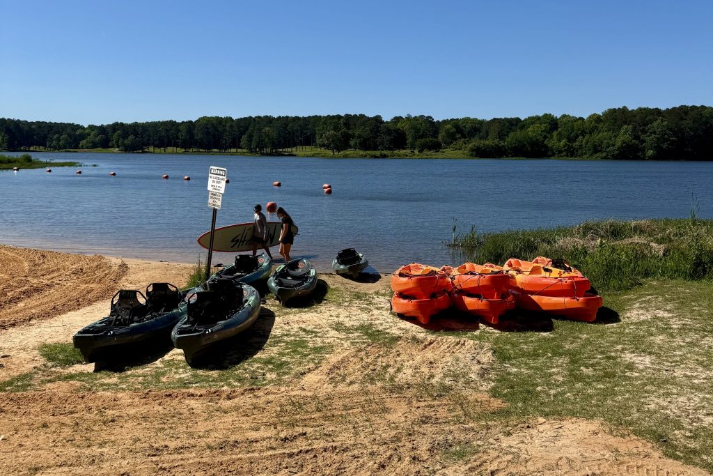 Several kayaks and paddleboards are on a grassy shore by a lake, with two people standing near a sign and trees visible across the water.