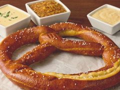 A large salted soft pretzel on parchment paper with three dipping sauces in square dishes: cheese, mustard, and a creamy dip.