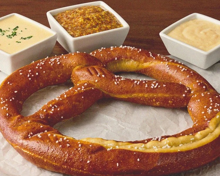 A large salted soft pretzel on parchment paper with three dipping sauces in square dishes: cheese, mustard, and a creamy dip.