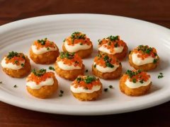 Ten bite-sized appetizers with a crispy base, topped with a dollop of cream, orange fish roe, and sprinkled with chopped herbs, arranged neatly on a white plate.