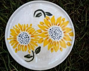 A round white plate with two yellow sunflowers and green leaves painted on it, placed on grass.