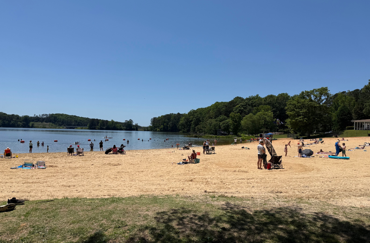 People relax and swim at a sandy beach by a lake on a clear, sunny day, with trees and grassy areas surrounding the shoreline—perfect for planning your beach day itinerary in Acworth.