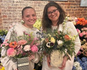 Two women standing indoors, smiling and holding flower arrangements in decorative containers, with a background of various colorful flowers and an exposed brick wall.