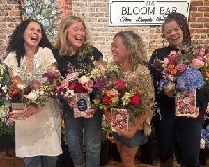 Four women stand together indoors, smiling and holding floral arrangements in decorative boxes at a shop called The Bloom Bar. A brick wall and flower display are in the background.