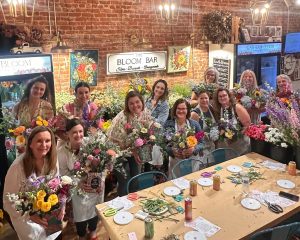 A group of women stand and sit around a table inside a flower shop, each holding colorful floral arrangements and smiling at the camera.