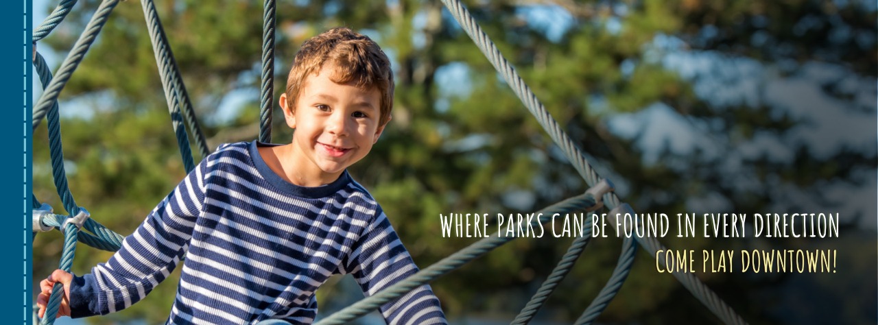 A young boy in a striped shirt climbs on playground ropes with trees in the background. Text reads, “Where parks can be found in every direction. Come play downtown!”.