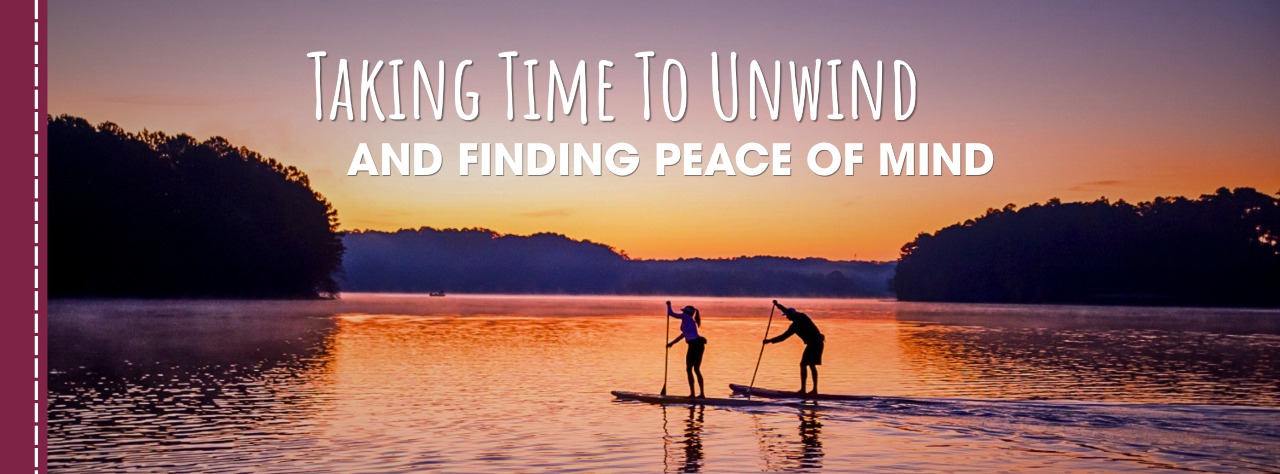 Two people paddleboarding on a calm lake at sunset with the text "Taking Time To Unwind and Finding Peace of Mind" displayed above them.
