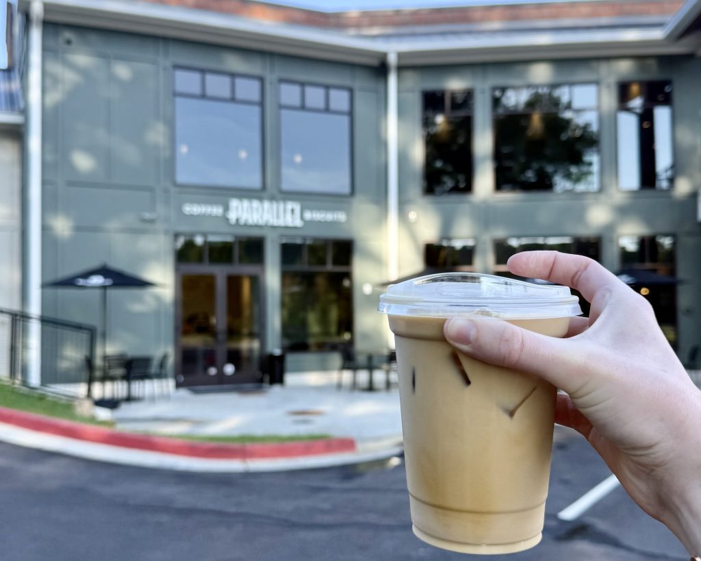 A hand holding a plastic cup of iced coffee in front of a modern coffee shop with 