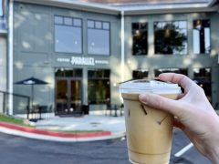 A hand holding a plastic cup of iced coffee in front of a modern coffee shop with "PARALLEL" on the sign above the entrance.