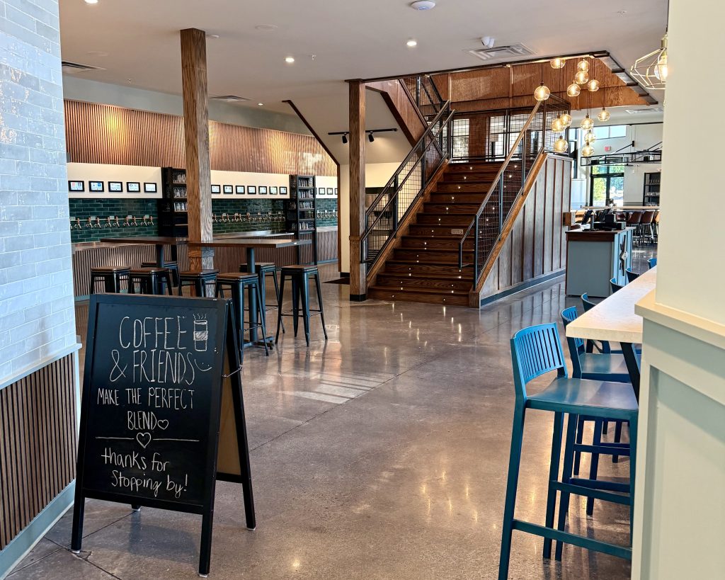 A modern café interior with wooden stairs, high tables and chairs, wall art, and a chalkboard sign that reads, 