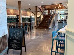 A modern café interior with wooden stairs, high tables and chairs, wall art, and a chalkboard sign that reads, "Coffee & friends make the perfect blend. Thanks for stopping by!.